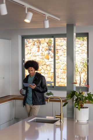 African american man standing using tablet drinking coffee in modern sunlit kitchen