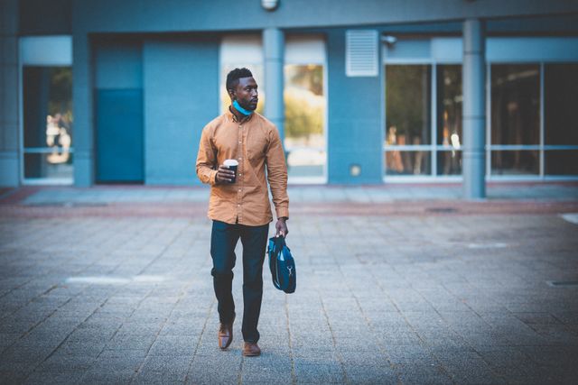 African American man wearing face mask walking in urban area holding coffee and bag. Suitable for themes related to modern lifestyle, urban living, professional life, pandemic safety, commuting, and health precautions.