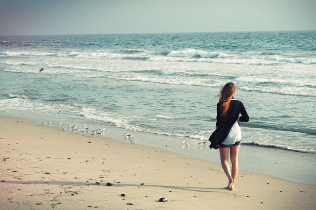 A young woman is walking barefoot along a sandy beach with gentle waves crashing in the background. She is dressed casually, enjoying a peaceful moment by the sea. This image conveys a sense of tranquility and relaxation, ideal for themes of freedom, vacation, and mindfulness. It can be used for travel promotions, lifestyle blogs, mental wellness articles, and advertisement related to beach resorts.