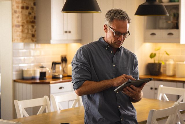 Middle-aged man wearing glasses using tablet in a modern kitchen. Ideal for themes related to technology use at home, remote work, casual lifestyle, and domestic settings. Can be used in articles about home technology, work-life balance, or lifestyle blogs.