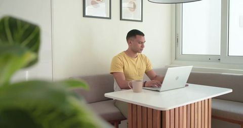 Young man working remotely at cozy minimalist dining nook with laptop, coffee and plant