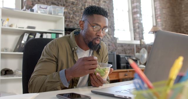 Young professional eating healthy salad while working at desk in bright modern office. Ideal for concepts of multitasking, healthy lifestyle at work, and balance between work and diet.