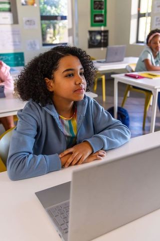 Thoughtful schoolgirl using laptop in bright classroom studying and focusing on lesson