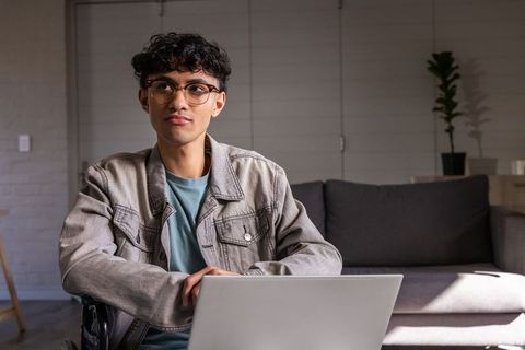 Asian man working on laptop in living room wearing light gray denim jacket and round glasses