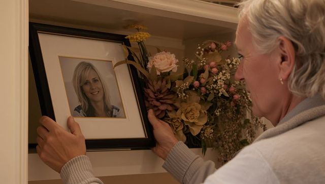 Senior woman arranging framed portrait on shelf with floral arrangement and warm light