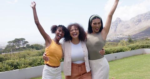 Three african american women laughing and embracing on backyard lawn with mountain view