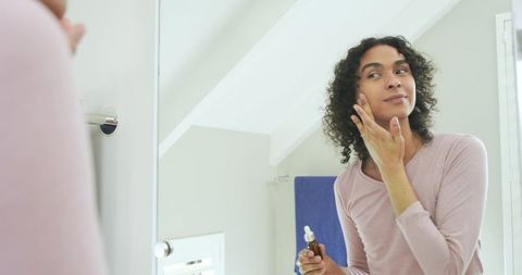 Nonbinary person applying facial serum at mirror in loft bathroom for skincare routine