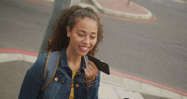 Young woman is outdoors on a sunny day, using her smartphone to send voice messages. Perfect for illustrating modern communication, technology in everyday life, social media use, or urban lifestyle.