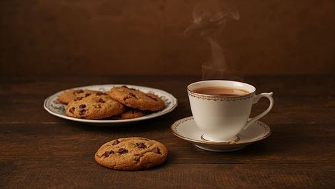 Steaming tea in gold-rim porcelain cup with chocolate chip cookies on rustic oak table