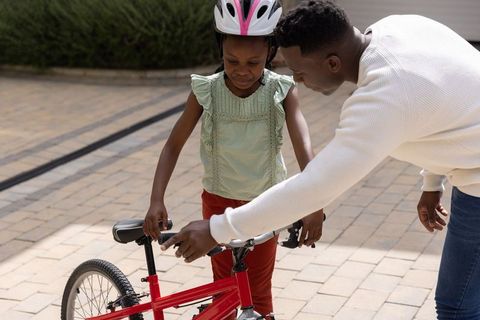 African american father helping daughter ride red bicycle on driveway