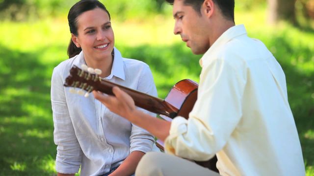 Video shows a man playing guitar for his girlfriend in a park, both wearing casual clothing and sitting on the grass. The scene captures a moment of joy and connection, suitable for use in promoting outdoors activities, dating apps, lifestyle blogs, or marketing campaigns focusing on romantic experiences.