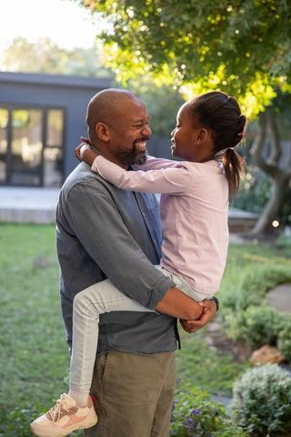 African american father lifting daughter in sunlit backyard embracing joyful family bond