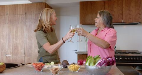 Two women are raising glasses of wine over a kitchen island filled with fresh vegetables. Enjoying a moment of camaraderie, this scene captures a joyful celebration within a warmly lit home. Useful for illustrating themes of friendship, special occasions, and joyful home gatherings in a contemporary lifestyle setting.