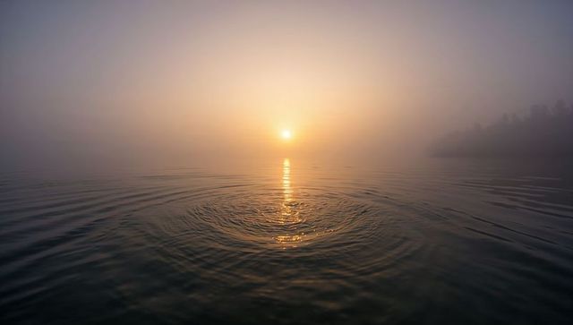 Misty sunrise over calm lake, sun casting golden reflection and concentric ripples
