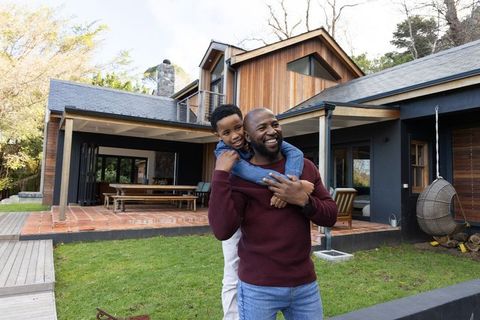 African american father carrying son on back near wooden house patio, smiling outdoors