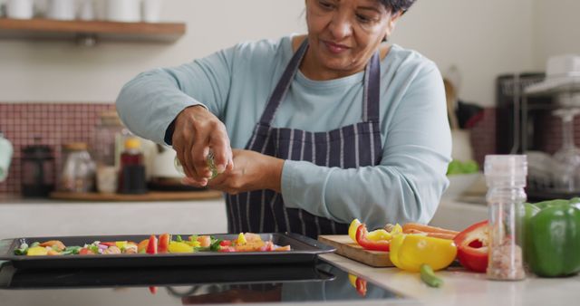 Elderly woman wearing an apron is preparing colorful vegetables on a baking tray in a kitchen. There are various vegetables like bell peppers and carrots on the counter. This can be used for topics on healthy eating, home cooking, senior lifestyle, and nutritious recipes.