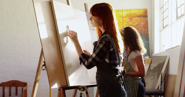 Female artists painting on canvases in a well-lit studio with large windows. Both wearing aprons, one with red hair in focus. Ideal for websites or articles about art, creativity, painting workshops, or collaborative artistic work.