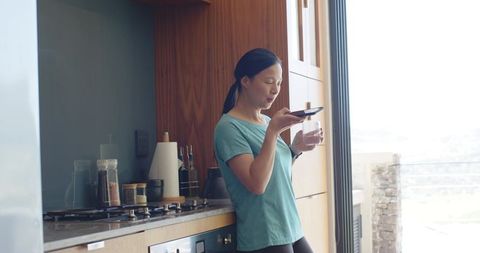 Asian woman speaking into smartphone while holding glass in sunlit modern kitchen