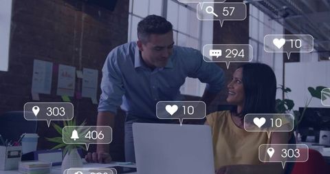 Manager Leaning Over Desk Guiding Colleague in Open-Plan Office with Social Notifications