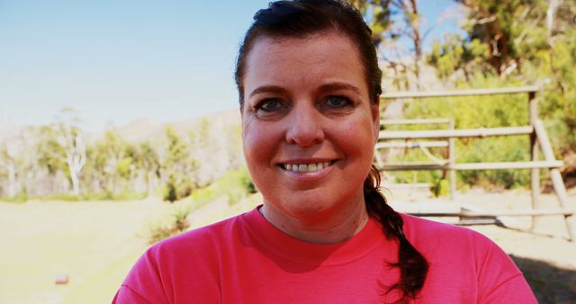 Woman standing outdoors with a bright smile, wearing a pink shirt. Trees and park elements in the background. Useful for concepts related to happiness, confidence, positivity, outdoor activities, and casual attire.