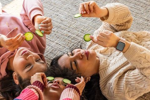 Diverse female family lying head-to-head holding cucumber slices for cozy at-home spa self-care