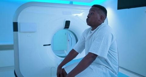 African american teen sitting on ct scanner table wearing gown and holding breath