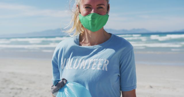 Portrait of caucasian woman wearing volunteer t shirt and face mask looking at camera. eco conservation volunteers, beach clean-up during coronavirus covid 19 pandemic.