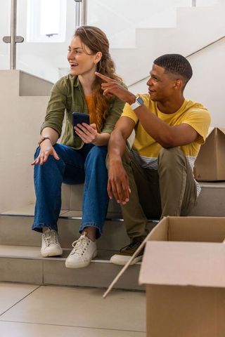 Laughing young couple unpacking boxes on stairs while pointing and holding smartphone