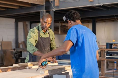 African american mentor training apprentice measuring wood on workbench woodworking shop