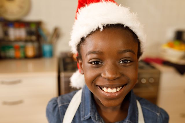 This image captures a joyful African-American girl wearing a Santa hat and smiling in a kitchen. Perfect for holiday-themed promotions, Christmas cards, festive advertisements, and family-oriented content. The warm and cheerful atmosphere makes it ideal for conveying holiday spirit and joy.