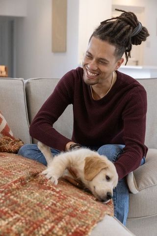 Smiling man with dreadlocks holding golden retriever puppy on cozy couch