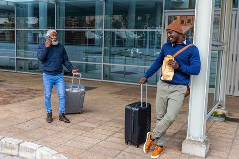 Two men waiting outside modern glass terminal with suitcases, chatting and eating snack