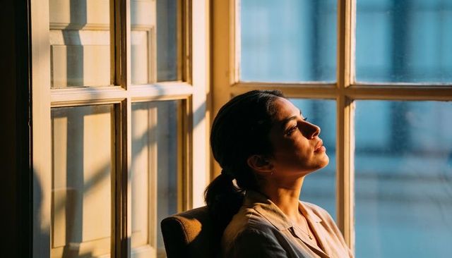 Woman soaking in golden sunlight at paned window, gazing contemplatively, minimalist interior