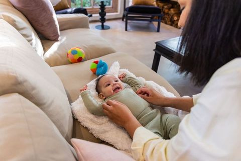 Smiling infant bonding with caregiver on plush sofa while adjusting light green knit romper