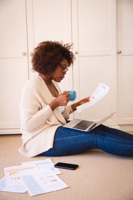 African American woman sitting on floor in living room, working on laptop while holding coffee cup and reviewing documents. Ideal for illustrating concepts of remote work, home office setup, multitasking, and modern work-life balance.
