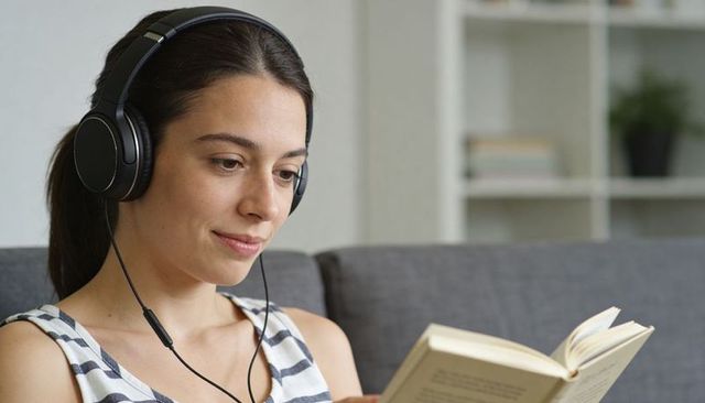 Young woman reading with headphones on gray sofa at home, cozy audiobook and study moment