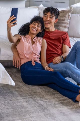Young couple taking selfie on cozy living room floor with floating heart graphics