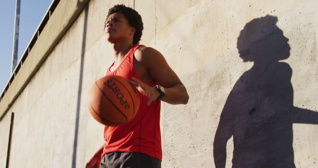 Young man in red tank top playing basketball in urban setting with concrete wall casting a shadow. Suitable for use in sports, fitness, youth activities, and urban lifestyle themes. Ideal for ads, blogs, and social media posts focusing on athleticism, outdoor sports, and energetic lifestyles.