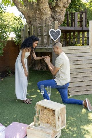Backyard proposal moment: man kneeling presenting ring to partner by rustic treehouse