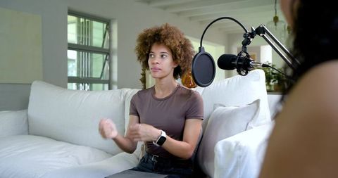 Young african american woman recording podcast on sofa speaking into studio microphone