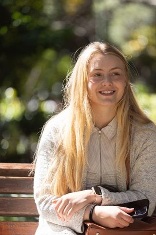 Smiling young woman sitting on wooden park bench holding leather bag and phone in sunlight