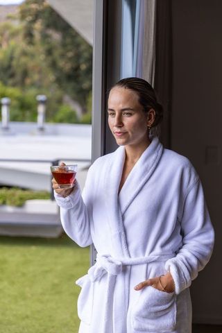 Woman in white robe standing at sliding door holding amber drink gazing outdoors serene morning
