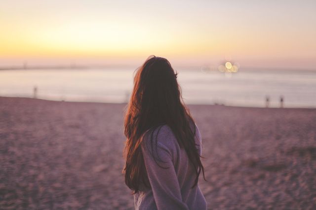 This image features a woman with long hair standing on a beach, watching the sunset create a pink hue in the sky and reflecting over the calm sea. Use this for themes related to relaxation, nature, peaceful moments, or contemplative solitude.