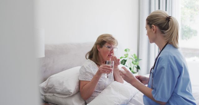 Nurse in blue scrubs aids elderly woman in comfortable home setting by providing pill and water. Suitable for use in articles and promotions about home healthcare, caregiving, elderly assistance, and the importance of medical support for senior citizens.