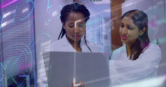 Two female healthcare professionals, wearing white coats, discussing information displayed on a laptop in a modern laboratory. The lab has a technological setup that includes digital interfaces reflected in the scene. This image can be used for topics related to technology in healthcare, teamwork in medical research, or scientific analysis in a clinical setting.