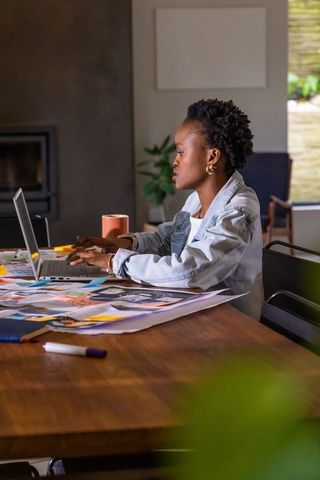 African american designer working on laptop in home office surrounded by printed posters