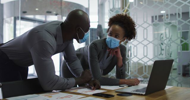 Diverse male and female business colleagues in face masks discussing, using laptop in office. business professional working in modern office during covid 19 coronavirus pandemic.