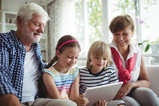 Grandparents and grandchildren sitting together on a couch, engaging with a digital tablet. This image is perfect for illustrating family bonding, technology use across generations, and happy family moments. Ideal for use in advertisements, educational materials, and articles about family life, technology in education, and intergenerational relationships.