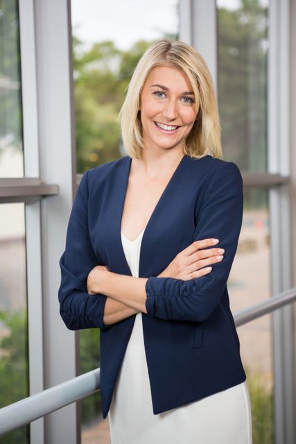 Professional woman with blonde hair stands confidently with arms crossed in a modern office. Ideal for corporate, leadership, teamwork, business presentations, and diversity in the workplace themes.
