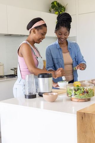 African mother and teenage daughter cooking together at modern kitchen island, smiling